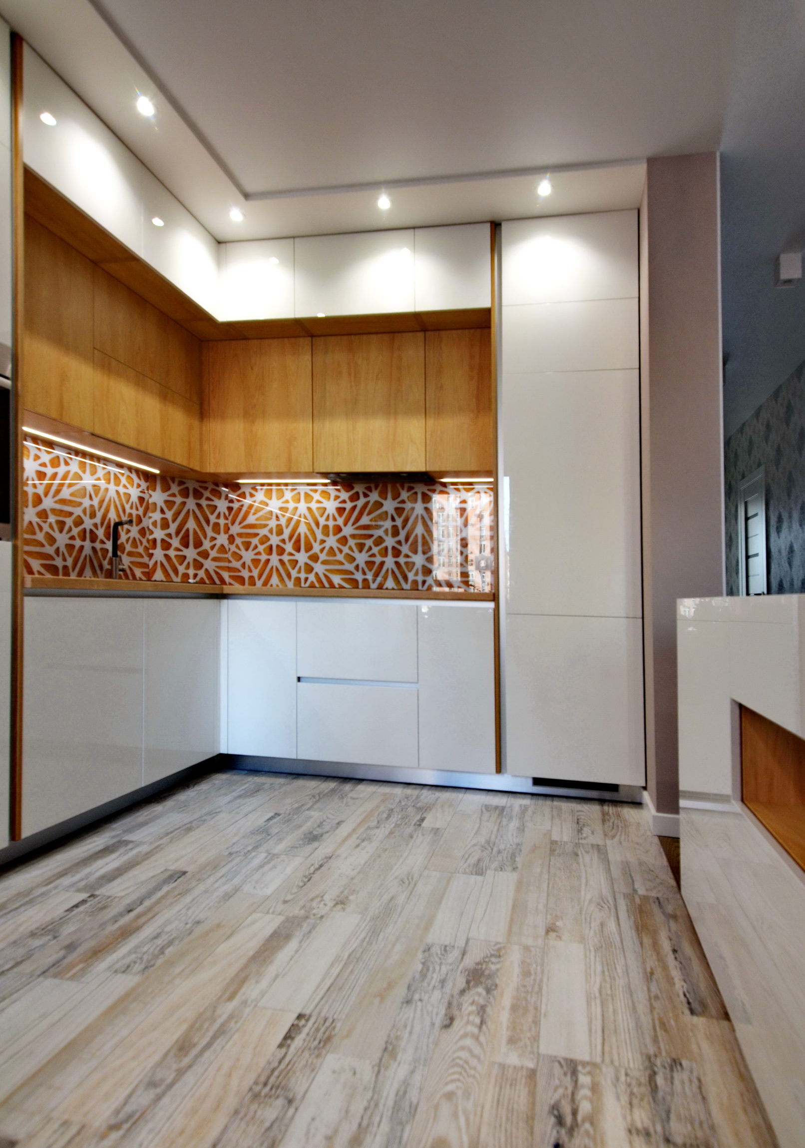 Сучасна кутова кухня з білими глянцевими фасадами, дерев'яними верхніми шафами та акцентним скляним скіналі з помаранчевим графічним візерунком